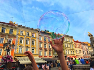 Several people enjoy a vibrant urban scene, with a focus on a large, translucent soap bubble against a backdrop of colorful old buildings. Hands reach up playfully towards the bubble. The atmosphere suggests a lively and cheerful gathering in a public square.