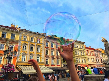 Several people enjoy a vibrant urban scene, with a focus on a large, translucent soap bubble against a backdrop of colorful old buildings. Hands reach up playfully towards the bubble. The atmosphere suggests a lively and cheerful gathering in a public square.