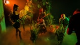 A group of people in a dimly lit, jungle-like environment with lush green foliage. They appear to be exploring or preparing for an adventure. The lighting creates deep shadows and a mysterious atmosphere with a warm orange and green tint.