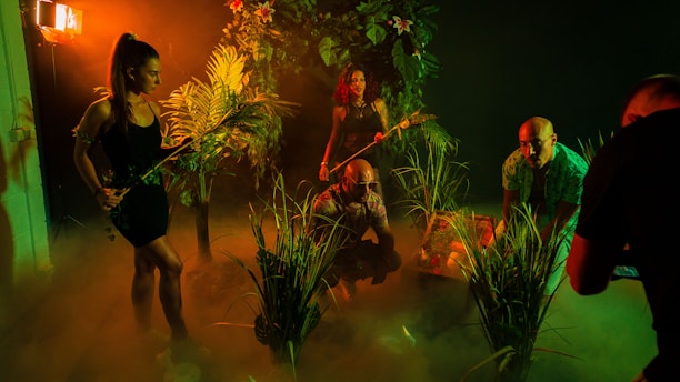 A group of people in a dimly lit, jungle-like environment with lush green foliage. They appear to be exploring or preparing for an adventure. The lighting creates deep shadows and a mysterious atmosphere with a warm orange and green tint.