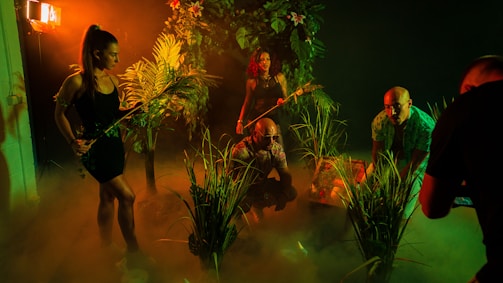A group of people in a dimly lit, jungle-like environment with lush green foliage. They appear to be exploring or preparing for an adventure. The lighting creates deep shadows and a mysterious atmosphere with a warm orange and green tint.