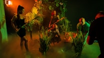 A group of people in a dimly lit, jungle-like environment with lush green foliage. They appear to be exploring or preparing for an adventure. The lighting creates deep shadows and a mysterious atmosphere with a warm orange and green tint.