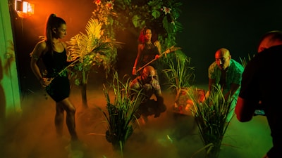 A group of people in a dimly lit, jungle-like environment with lush green foliage. They appear to be exploring or preparing for an adventure. The lighting creates deep shadows and a mysterious atmosphere with a warm orange and green tint.