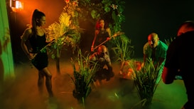 A group of people in a dimly lit, jungle-like environment with lush green foliage. They appear to be exploring or preparing for an adventure. The lighting creates deep shadows and a mysterious atmosphere with a warm orange and green tint.