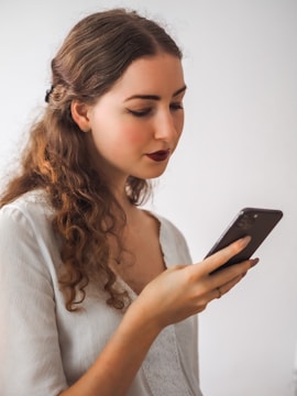 A young woman with long curly hair and a gentle expression is looking at a smartphone in her hand. She is wearing a white blouse with lace detailing and has dark lipstick. The background is plain and minimalistic, highlighting her relaxed focus on the device.