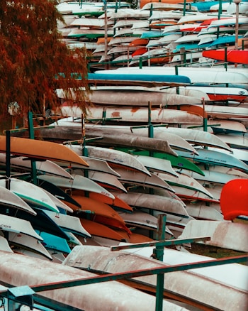 A large collection of kayaks and canoes stacked horizontally on metal racks, variously colored with shades of blue, green, red, and orange. The scene appears to be outdoors near a body of water, with trees partially visible on the left side.