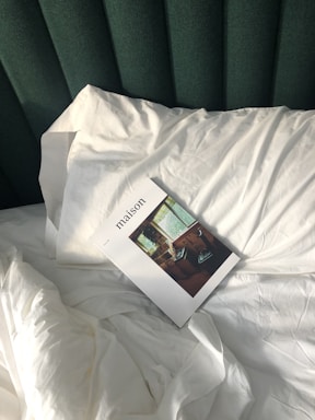A magazine titled 'maison' rests on a neatly made bed with white linens and a green upholstered headboard. The image on the magazine cover shows an interior scene with a window and a chair.