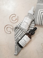 A minimalist arrangement featuring two dropper bottles with 'The Ordinary' labels placed on a marble surface. Accompanying the bottles, a pair of gold hoop earrings are set beside a black-and-white striped fabric. The aesthetic is clean and contemporary, with an emphasis on skincare and fashion accessories.