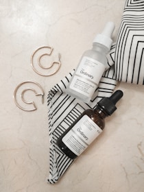 A minimalist arrangement featuring two dropper bottles with 'The Ordinary' labels placed on a marble surface. Accompanying the bottles, a pair of gold hoop earrings are set beside a black-and-white striped fabric. The aesthetic is clean and contemporary, with an emphasis on skincare and fashion accessories.