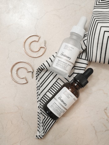 A minimalist arrangement featuring two dropper bottles with 'The Ordinary' labels placed on a marble surface. Accompanying the bottles, a pair of gold hoop earrings are set beside a black-and-white striped fabric. The aesthetic is clean and contemporary, with an emphasis on skincare and fashion accessories.