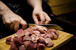 Hands are cutting raw pieces of meat on a wooden cutting board with a sharp knife.