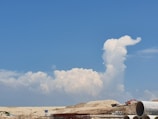 A construction site with heavy machinery and stacks of large concrete pipes in the foreground. In the background, a large, fluffy white cloud formation rises against a vibrant blue sky, resembling a plume or a figure.