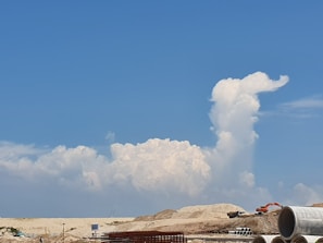A construction site with heavy machinery and stacks of large concrete pipes in the foreground. In the background, a large, fluffy white cloud formation rises against a vibrant blue sky, resembling a plume or a figure.