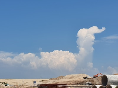 A construction site with heavy machinery and stacks of large concrete pipes in the foreground. In the background, a large, fluffy white cloud formation rises against a vibrant blue sky, resembling a plume or a figure.