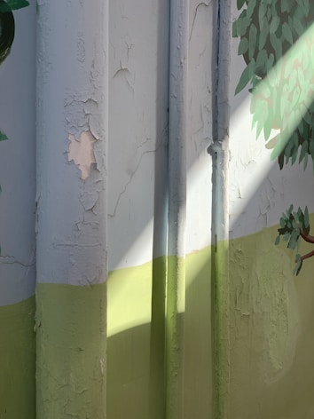 Peeling paint on a wall with a design featuring pipes and a painted tree. Light casts shadows across the surface, highlighting the texture of the flaking paint. The colors divide the wall with a contrasting lower green section and upper pale section.