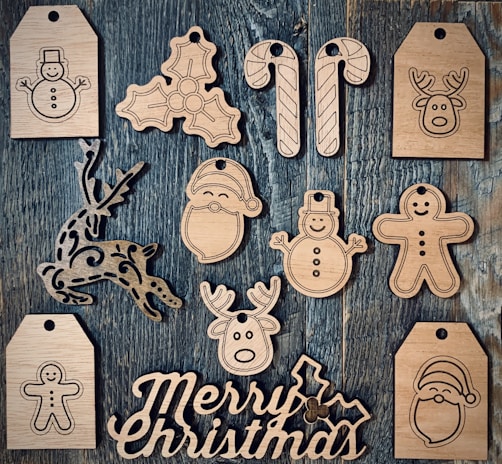 Wooden cutouts featuring various Christmas-themed shapes, including snowmen, candy canes, reindeer, holly leaves, a Santa Claus face, and a gingerbread man, arranged on a textured wooden surface. The words 'Merry Christmas' are also carved in wood.
