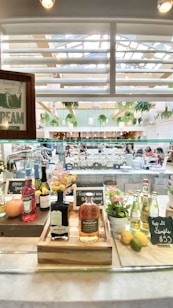A well-lit indoor space with a variety of bottles on display, including Aperol and Redemption Rye. There are decorative flowers and citrus fruits placed on a counter. In the background, people are seated in what appears to be a lively cafe or restaurant environment with a number of hanging plants and modern decor.