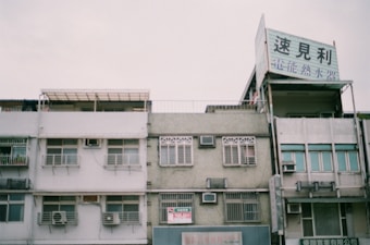 The image features a row of older apartment buildings, characterized by their boxy architecture and drab, weathered exteriors. Several of the windows have air conditioning units installed, and some of the balconies appear to have been enclosed. A large sign in Chinese text is prominently displayed on the rooftop of one building. The sky above is overcast, giving the scene a muted, slightly melancholic feel.