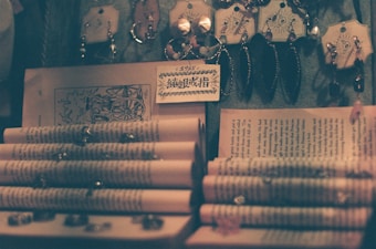 A collection of jewelry items hangs neatly from hooks, placed above open books with visible text. The scene is dimly lit, casting a soft, warm glow that enhances the vintage and antique appearance of both the jewelry and the books. Some pages of the books are curled, and a card with intricate writing is displayed prominently.