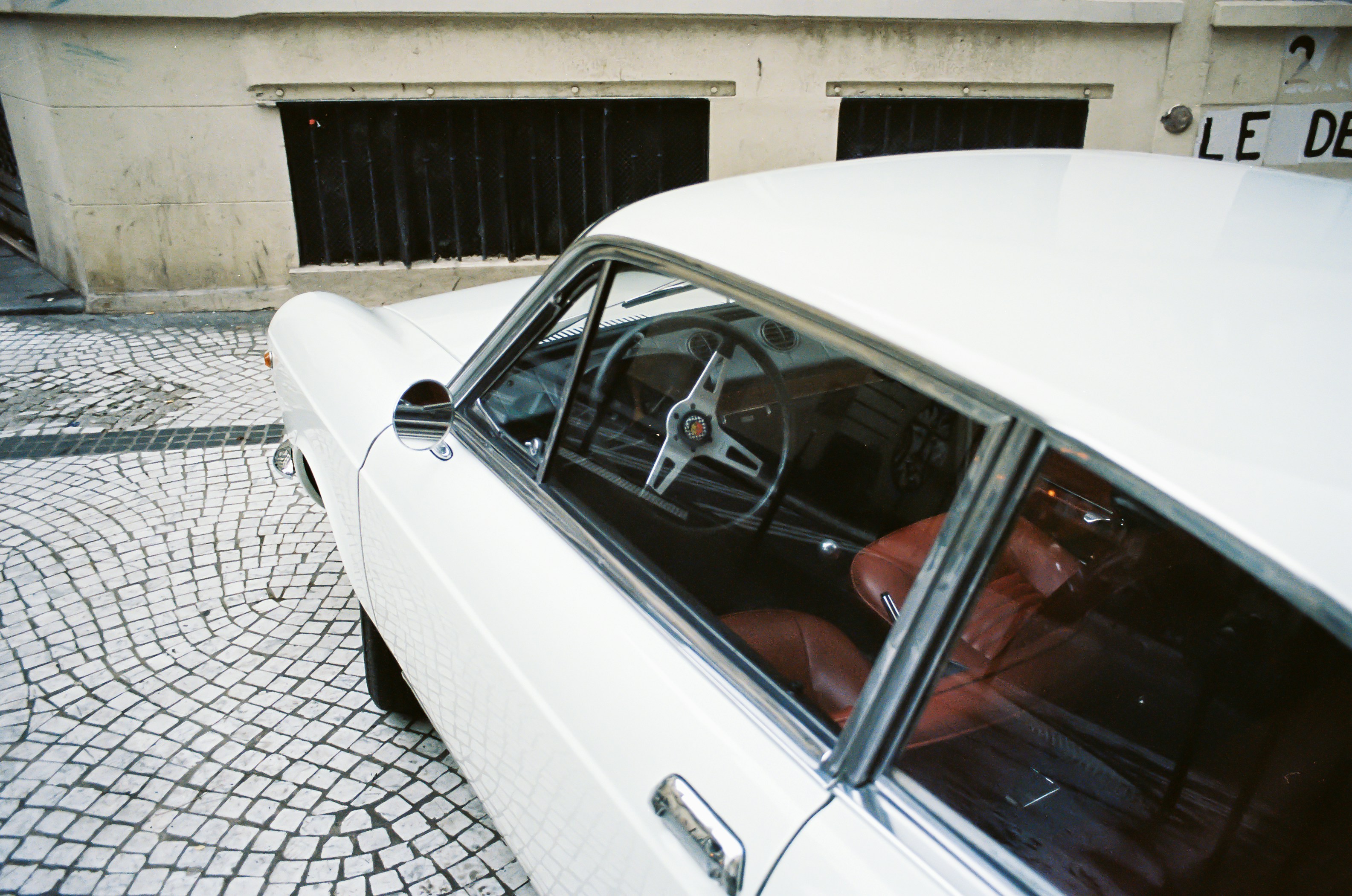 A white vintage car with a warm leather interior stands on pale stone pavement.