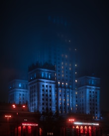 A grand building illuminated with blue and red lights stands prominently in the night, partially obscured by a mist. The lower part of the building features a sign that reads 'Teatr Dramatyczny' highlighted in red lighting. The architectural style appears monumental with tall columns and detailed cornices.