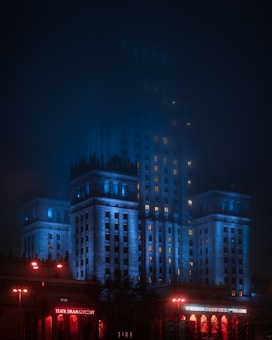 A grand building illuminated with blue and red lights stands prominently in the night, partially obscured by a mist. The lower part of the building features a sign that reads 'Teatr Dramatyczny' highlighted in red lighting. The architectural style appears monumental with tall columns and detailed cornices.