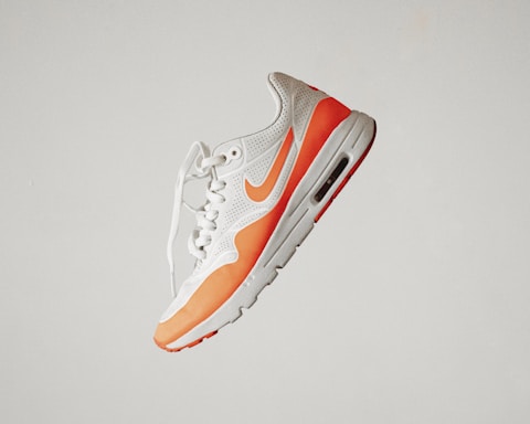 A stylish athletic sneaker featuring a white and orange color scheme, with perforated detailing and a visible air cushion in the sole. The shoe is positioned against a plain background, emphasizing its sleek and modern design.