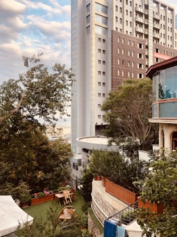 A tall, modern high-rise building with a mix of reflective and dark panels is situated next to lush greenery and trees. In the foreground, there is a garden area with wooden outdoor furniture, potted plants, and a decorative stone wall. A partial view of a smaller, elegant building with large windows is also visible. The sky is partly cloudy, casting soft light over the scene.