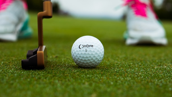 A golf ball labeled 'OnCore 1' is positioned on a lush green grass surface, with a brown and black putter poised nearby. In the background, blurred brightly colored athletic shoes suggest a person is preparing to play.