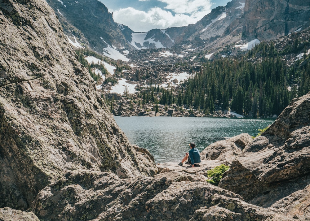 personne assise sur un rocher près d’un lac et d’une chaîne de montagnes