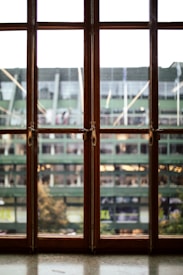 A wooden window with multiple panes overlooks a blurred exterior view of a green structure, possibly a stadium, with flags and banners visible in the distance.