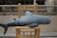 A plush shark toy is resting on a wooden chair located outdoors. The background is blurred, highlighting the soft texture and blue-gray color of the toy.