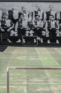 A vintage black and white photograph of a men's soccer team is displayed on a large mural. Below the mural is an empty sports court with faded green paint and a metal goalpost.