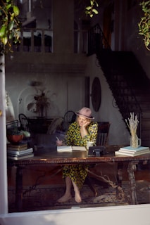 A woman wearing a floral dress and a hat is sitting barefoot at a wooden table in a warmly lit room. She appears to be writing in a notebook, with her head resting on one hand. The setting includes an eclectic mix of furniture, books, and decorative plants. The room has elegant architectural details including a staircase and large window, through which this scene is visible.