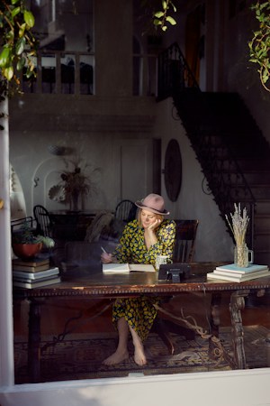 A woman wearing a floral dress and a hat is sitting barefoot at a wooden table in a warmly lit room. She appears to be writing in a notebook, with her head resting on one hand. The setting includes an eclectic mix of furniture, books, and decorative plants. The room has elegant architectural details including a staircase and large window, through which this scene is visible.