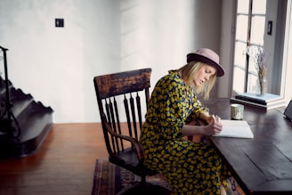 A person in a patterned dress and hat is seated at a wooden table, writing in a notebook. The room is well-lit with natural light coming from a window next to the table. Decor items, such as a cup and a vase with dried flowers, are placed on the table, adding a cozy feel to the space.
