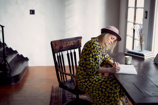 A person in a patterned dress and hat is seated at a wooden table, writing in a notebook. The room is well-lit with natural light coming from a window next to the table. Decor items, such as a cup and a vase with dried flowers, are placed on the table, adding a cozy feel to the space.