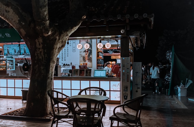 A small, dimly lit food stall situated outdoors, surrounded by trees. The stall has a wooden structure with illuminated signage in Chinese. Inside, a food vendor is attending to customers, with various items and a menu visible. A few customers are gathered around, and a table with chairs is placed outside under a tree.