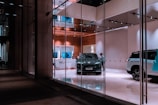 A modern showroom with glass walls features a sleek, dark-colored car prominently displayed in the center. Several people are present, possibly staff or customers, examining the vehicle. The interior has a sophisticated design with wooden elements and a clean, polished floor. Bright lighting highlights the cars and gives the space a luxurious feel.