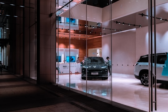 A modern showroom with glass walls features a sleek, dark-colored car prominently displayed in the center. Several people are present, possibly staff or customers, examining the vehicle. The interior has a sophisticated design with wooden elements and a clean, polished floor. Bright lighting highlights the cars and gives the space a luxurious feel.