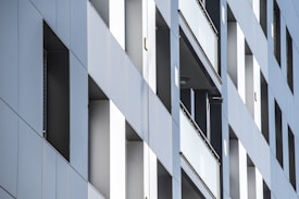 A modern building facade with a sleek, geometric design. The structure features large vertical rectangular windows and panels with a reflective surface, creating a minimalist and contemporary look.