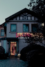 The exterior of a cafe and bar named 'La Luna' is visible, with a prominent neon sign displaying the name. The building is styled with dark trim and a star-shaped decoration above the second floor windows. A person can be seen through the glass door, and the surrounding area includes manicured bushes and a patterned pathway leading to the entrance.