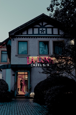 The exterior of a cafe and bar named 'La Luna' is visible, with a prominent neon sign displaying the name. The building is styled with dark trim and a star-shaped decoration above the second floor windows. A person can be seen through the glass door, and the surrounding area includes manicured bushes and a patterned pathway leading to the entrance.