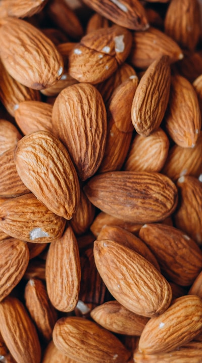 A close-up view of many almonds with their textured brown shells. The almonds are densely packed, filling the entire frame with a warm and earthy tone.