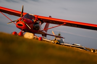 A vibrant red small aircraft is parked on a grassy field with other planes in the background. The plane is tilted slightly, with the nose and propeller prominently in view. The warm lighting suggests a sunset or sunrise atmosphere.