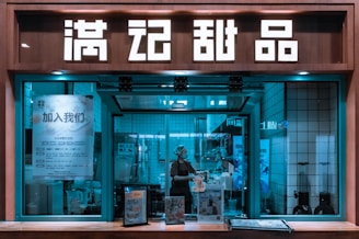 A storefront with large illuminated signs in an Asian script displayed above a window. Through the window, a person wearing a hairnet and mask is working, possibly preparing food. The interior has a tiled wall with various posters and a menu. There are also advertisements or displays on the counter in front of the window.