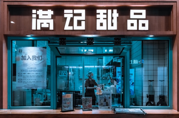A storefront with large illuminated signs in an Asian script displayed above a window. Through the window, a person wearing a hairnet and mask is working, possibly preparing food. The interior has a tiled wall with various posters and a menu. There are also advertisements or displays on the counter in front of the window.