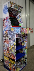 An arcade cabinet covered in colorful comic book graphics, including characters and scenes from various comic books. The cabinet has a joystick and buttons, and it is situated against a wall in a room with a concrete floor.