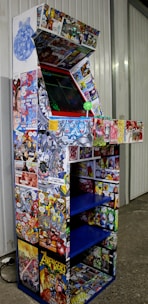 An arcade cabinet covered in colorful comic book graphics, including characters and scenes from various comic books. The cabinet has a joystick and buttons, and it is situated against a wall in a room with a concrete floor.