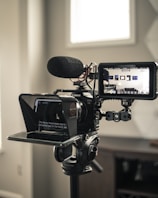 A professional video camera setup includes a large camera mounted on a tripod, equipped with a teleprompter and a monitor. The camera has a microphone attached, and the monitor displays a live feed of the room. The environment appears to be well-lit with natural light coming through a window.